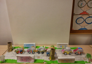 Prace dzieci - makieta wykonana z pudełek i papieru, przedstawiająca fragment ulicy: jezdnię, przejście dla pieszych, chodnik, budynki, drzewa, znaki drogowe oraz składane z papieru samochody